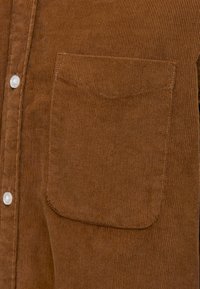 Brun manchesterskjorta med vertikala räfflor, en ficka med lapp och vita knappar längs framknäppningen.