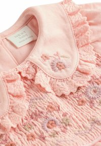 Vaaleanpunainen vauvan mekko, jossa on kirjailtuja kukkakuvioita, röyhelökaulus ja teksturoitu smokkityyli. Tunnuksessa lukee "NEXT baby, valmistettu Intiassa."