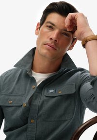 Man met donker haar die zijn hoofd op zijn hand steunt, gekleed in een grijs overhemd met knopen en omhoog geslagen kraag, met een bruine polsband om, zittend op een houten stoel.