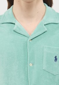 Camicia button-up in terry cloth verde chiaro con colletto, tasca frontale con logo navy e una texture morbida.