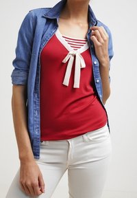 Mujer vestida con una blusa roja con un lazo blanco, detalles a rayas, una camisa de mezclilla azul con las mangas arremangadas y pantalones blancos.