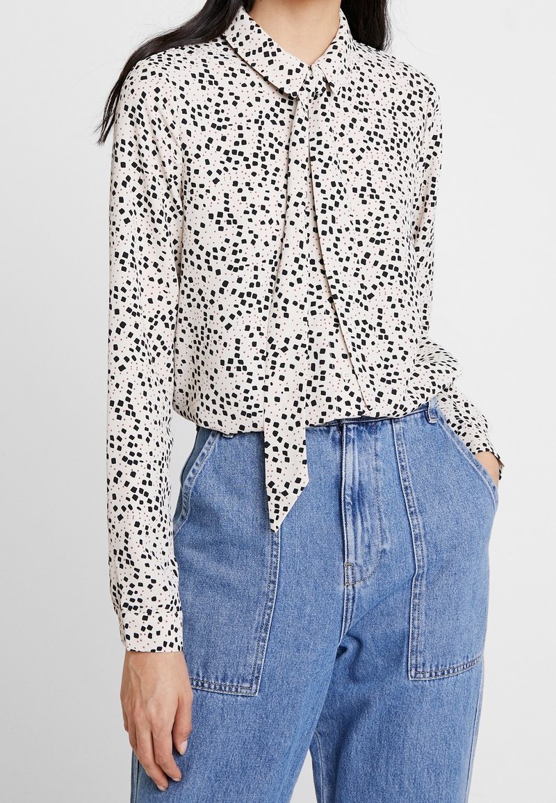 Femme portant un chemisier blanc à manches longues avec des taches noires et un nœud à l'avant, associé à un pantalon en denim bleu taille haute avec de grandes poches.