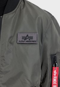 Olijfgroene bomberjack met een gladde textuur, voorzien van een grijs stoffen patch en een rood label met de tekst "REMOVE BEFORE FLIGHT."
