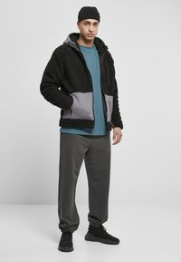 Jachetă din fleece neagră și gri cu glugă, închidere cu fermoar și buzunare laterale. Asortată cu o cămașă albastră și pantaloni gri, purtată cu adidași negri.