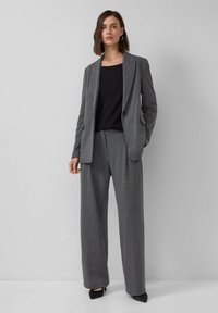 Grauer Pinstripes-Anzug mit einem taillierten Sakko und weiten Hosen, kombiniert mit einem schwarzen Oberteil. Besitzt elegante Linien und subtile Textur.