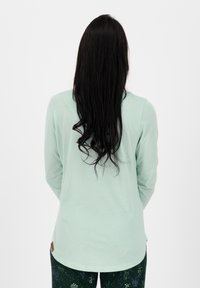 Mintgroene langekoot met een ronde zoom, zachte textuur en een klein bruin labeldetail aan de linkerkant. Draagt met een gebloemde donkere broek.