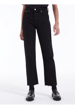 Personne portant un jean noir taille haute à jambes droites avec des ourlets effilochés, des chaussettes noires et des chaussures slip-on noires sur fond blanc.