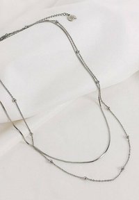 Collana in argento con design a catena multipla, una barra curva liscia e perline tonde distribuite lungo la catena, poggiata su una superficie bianca.