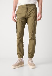 Olijfgroene cargobroek met een elastische tailleband, ritszakken aan de zijkant en elastische boorden. Draag met beige suède sneakers.