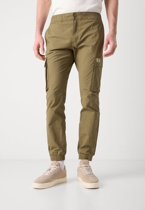 Pantalones cargo verde oliva con cintura elástica, bolsillos laterales con cremallera y puños elásticos. Llevados con zapatillas de ante beige.