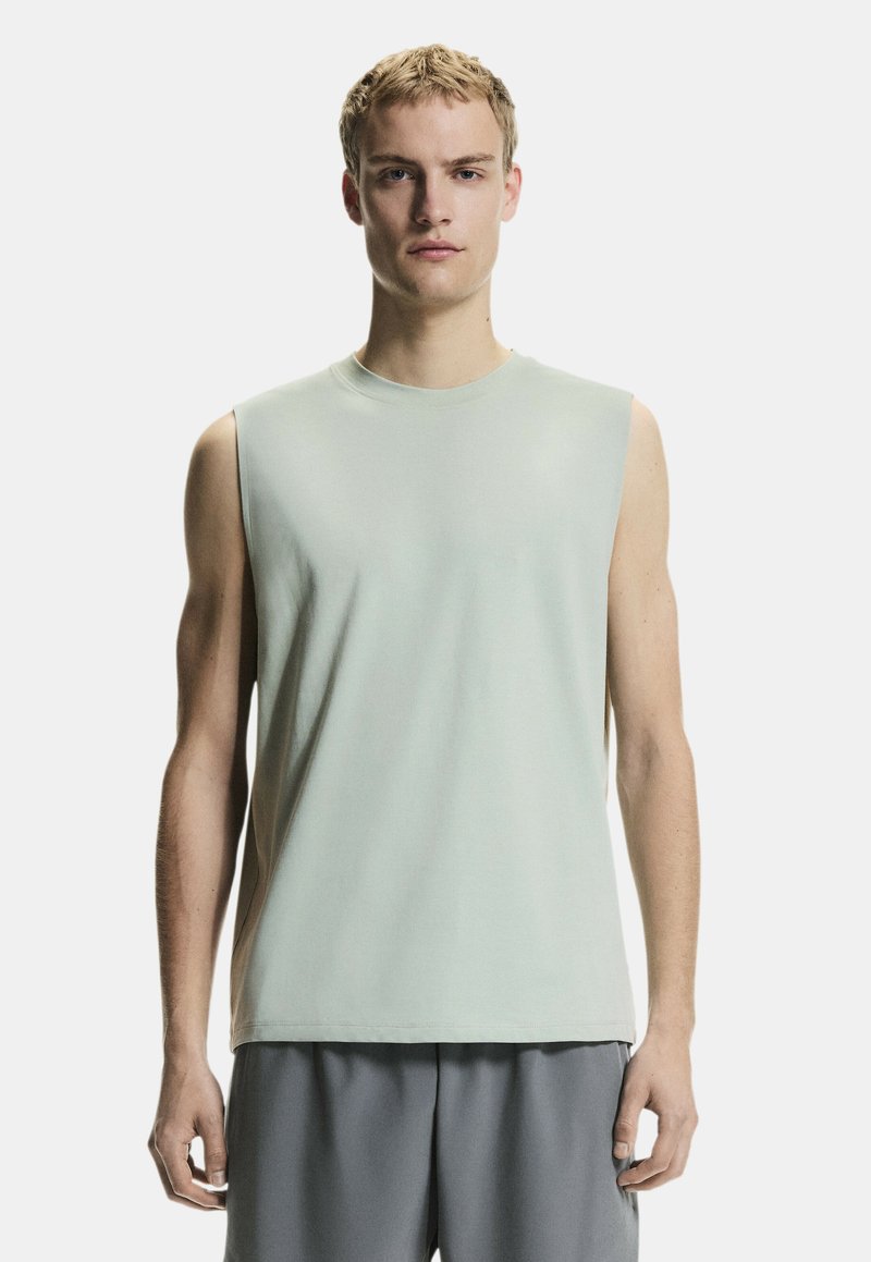 Jeune homme portant un débardeur gris clair et un pantalon ample gris, debout devant un fond blanc uni.