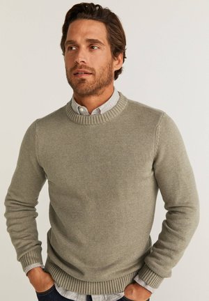 Grauer Strickpullover mit geripptem Ausschnitt und Bündchen, über einem hellen, kragenlosen Hemd getragen. Strukturierter Stoff, lässiger Schnitt.