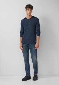 Dunkelblauer Strickpullover mit strukturiertem Design, Rundhalsausschnitt und langen Ärmeln. Kombiniert mit hellblauen Jeans und schwarzen Stiefeln.