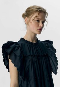 Schwarzes Kleid mit großen, gewellten Rüschensäumen, einer gerafften Brustpartie und einem hohen Rundhalsausschnitt, aus leichtem Stoff gefertigt.