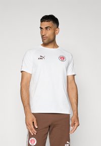 Bijela kratka majica s kratkim rukavima i okruglim izrezom, sadrži crveni kružni logo na prsima, te smeđi tekst i detalje. Mekana tkanina.