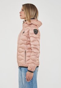 Donna con giacca a cappuccio trapuntata rosa e jeans blu in profilo contro uno sfondo bianco.