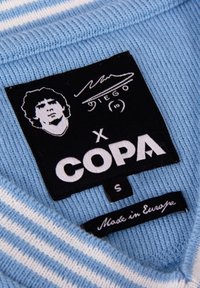 Tela azul claro a rayas con una etiqueta negra que presenta un logo blanco que dice "COPA." Incluye una firma y una etiqueta que dice "Hecho en Europa." Talla S.