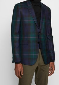 Geruite blazer in groen, marineblauw en zwart met een gestructureerd ontwerp, met notch revers en twee voorzakken. Draag comfortabel over een zwarte coltrui.