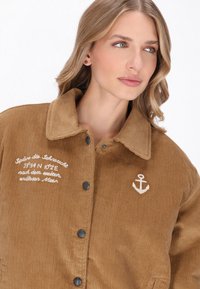 Barna kordbársony dzseki nyakkal, fekete gombokkal, hímzett szöveggel és horgony logóval a mellkason. Puha tapintású és függőleges bordázattal.