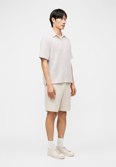 Νεαρός άνδρας που στέκεται πλαγίως φορώντας ανοιχτό μπεζ κοντομάνικο πουκάμισο με γιακά, ταιριαστά σορτς, λευκές κάλτσες και μπεζ αθλητικά παπούτσια.