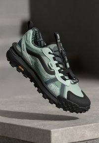 Zielone i czarne sneakersy z teksturowaną cholewką, podeszwą Vibram i oznakowaniem GORE-TEX. Charakteryzują się solidnym designem i kontrastującymi akcentami.