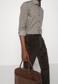 Bruine corduroy broek gecombineerd met een aansluitend shirt met een wervelend patroon in bruin, geel en blauw, vastgehouden met een bruine leren tas.