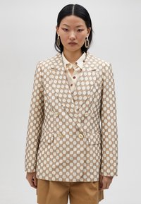 Dubbelknäppt blazer i beige och guld rutmönster; har klassiska slag, två framlommor och ljusa knappar. Kombinerad med en prickig skjorta.