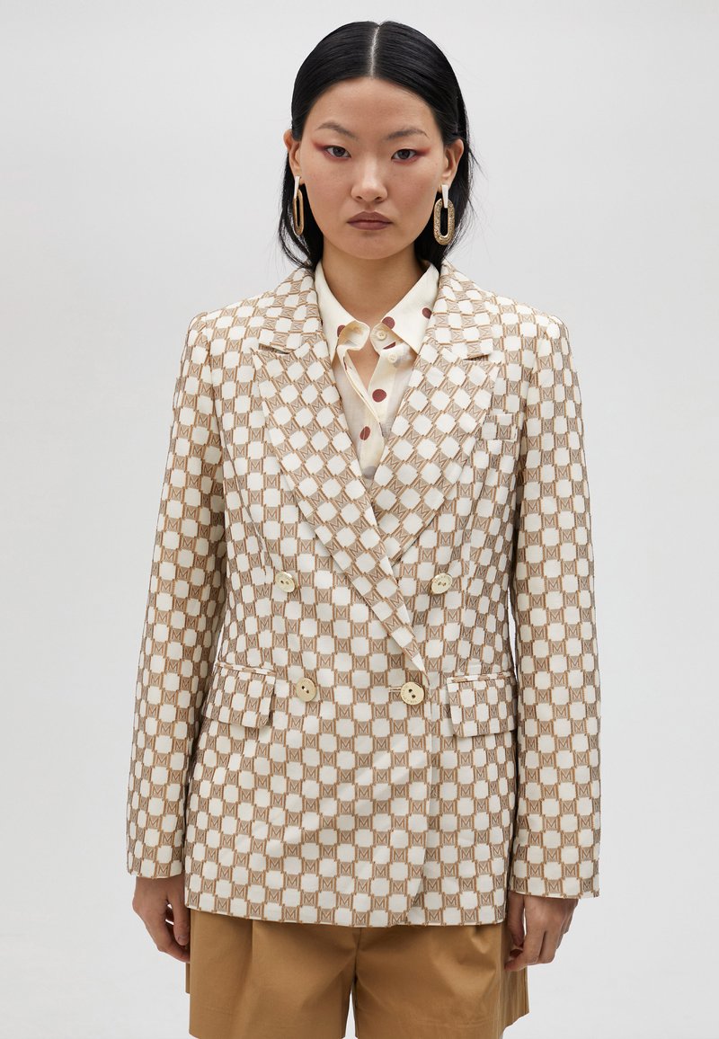 Dubbelknäppt blazer i beige och guld rutmönster; har klassiska slag, två framlommor och ljusa knappar. Kombinerad med en prickig skjorta.