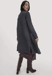 Langer, dunkelgrauer Cardigan aus Wollmischung, mit seitlichen Schlitzen, Taschen und lässiger Passform, kombiniert mit einem lila Rock und braunen kniehohen Boots.