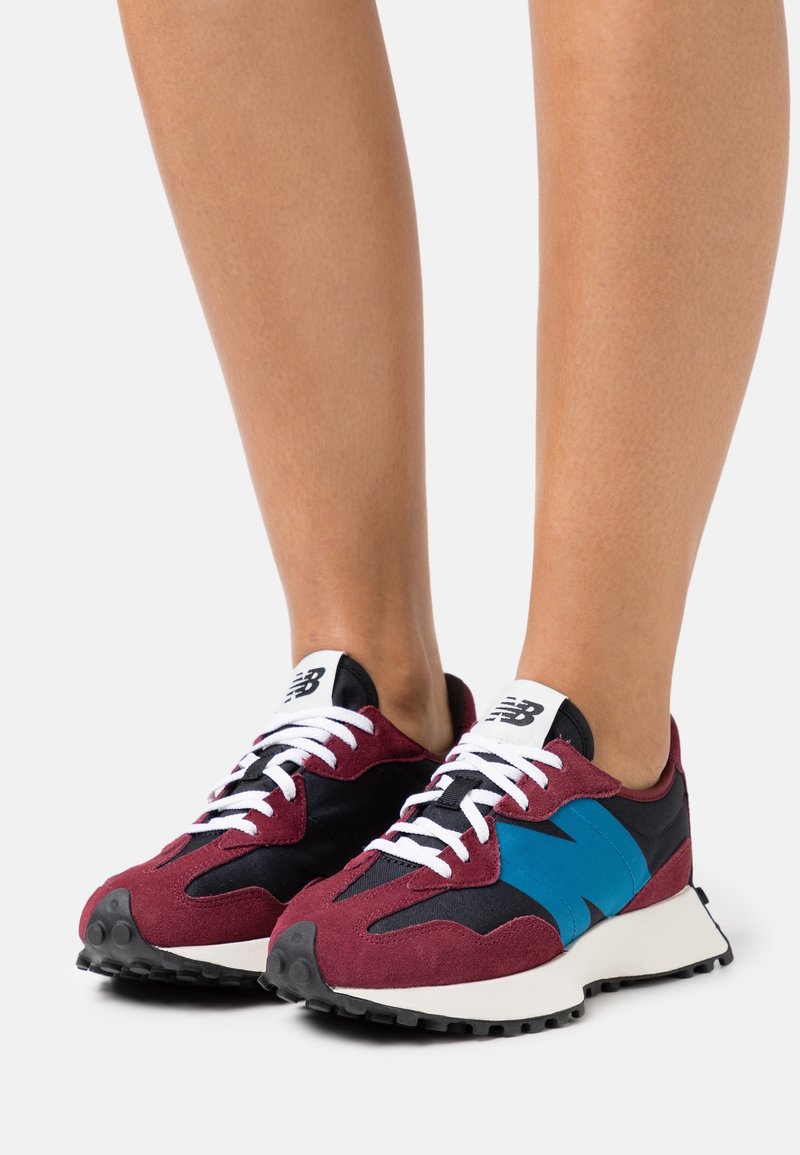 Paire de baskets New Balance bordeaux, noires et bleues avec des lacets blancs portées pieds nus sur un fond blanc.