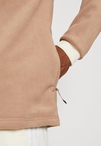 Fleece-pullover i ljusbeige med texturerat tyg, utrustad med en sidoficka med dragkedja och kontrasterande vita muddar.