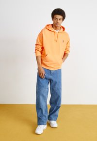 Oranje katoenen hoodie met buidelzak en koordkap, gecombineerd met lichtblauwe wijde denim jeans en witte sneakers.
