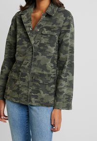 Green camo jacket with a relaxed shape, long sleeves, two front pockets, and snap button closure. Made of sturdy cotton fabric.