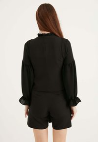 Schwarzes Outfit mit einem engen Oberteil mit Rüschensaum und langen Puffärmeln, kombiniert mit maßgeschneiderten Shorts aus demselben Material.