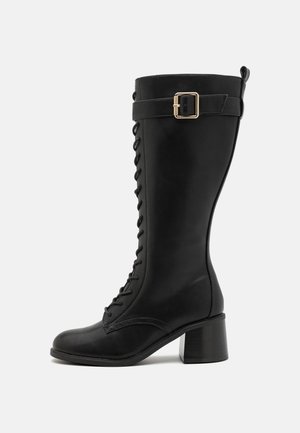 Zwarte leren kniehoog laars met een vierkante neus, vetersluiting aan de voorkant en een gespsluiting aan de zijkant. Blokhak biedt stabiliteit en hoogte.