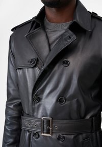 Black leather trench coat with double-breasted buttons, epaulets, and a belted waist. Features smooth texture and subtle sheen.