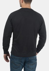 Sweat à col rond noir avec des manches raglan, des poignets et un ourlet côtelés. Tissu doux et texturé ; design uni. Porté avec un jeans en denim bleu.