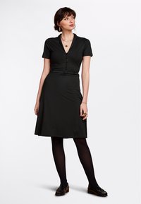Svart kortärmad klänning med V-hals, knapplist framtill, bälte i midjan och A-linjeskjort. Bärs med svarta strumpbyxor och loafers.
