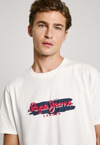 Camiseta de algodón blanca con un logotipo gráfico en rojo y azul que dice "Pepe Jeans LONDON" en el pecho con un estilo de escritura a mano. Mangas cortas.