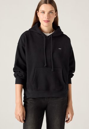 Frau trägt einen schwarzen Levi's-Hoodie mit Kordelzug und Fronttasche, kombiniert mit dunklen Jeans, steht vor einem schlichten Hintergrund.
