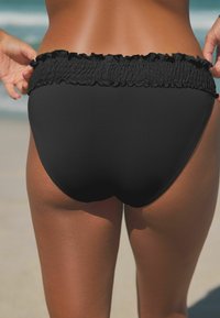 Bas de bikini noir avec une taille élastique froncée, texture lisse et design ajusté, sur fond de plage.