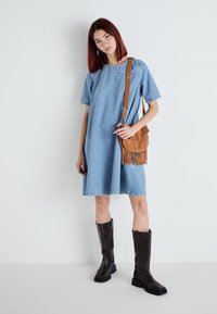 Džinsu kleita ar īsām piedurknēm un brīvu piegriezumu, apvienota ar brūnu zamšādas plecu somu ar franjām un ceļgalu augstiem melniem zābakiem.