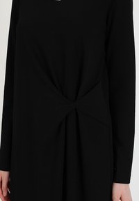 Vestido negro de manga larga hecho de una tela suave, con un detalle de nudo retorcido en la cintura y pliegues en la falda. Textura suave.