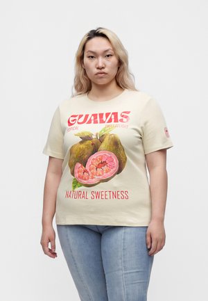 Frau mit langen blonden Haaren, die ein cremefarbenes T-Shirt trägt und Guavafrüchte sowie den Text „Guavas natürliche Süße“ zeigt, und hellblaue Jeans.
