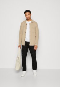 Veste chemise beige à boutons, t-shirt blanc, pantalon noir avec déchirures, baskets blanches, et un sac fourre-tout texturé blanc cassé.
