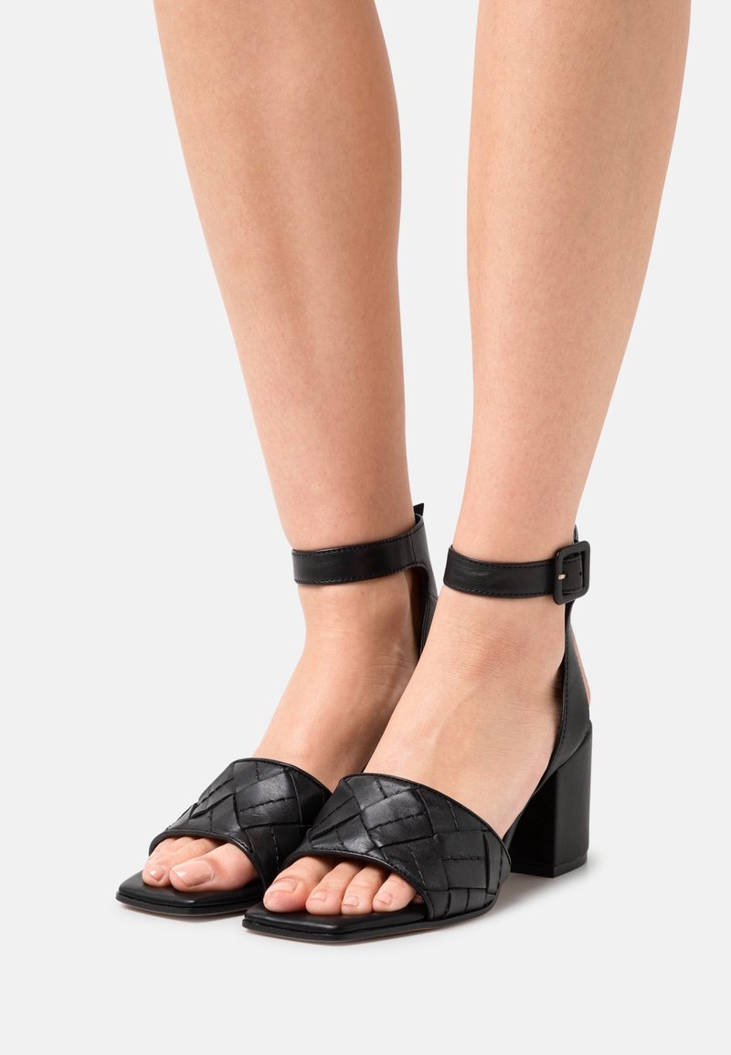 Pieds portant des sandales à talons hauts en cuir noir avec des brides tressées et des fermetures à boucle à la cheville sur un fond clair uni.