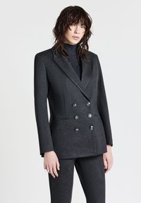 Blazer grigio scuro doppiopetto con finitura satinata, revers a lancia, sei bottoni e spalle strutturate. Abbinato a un dolcevita in maglia.