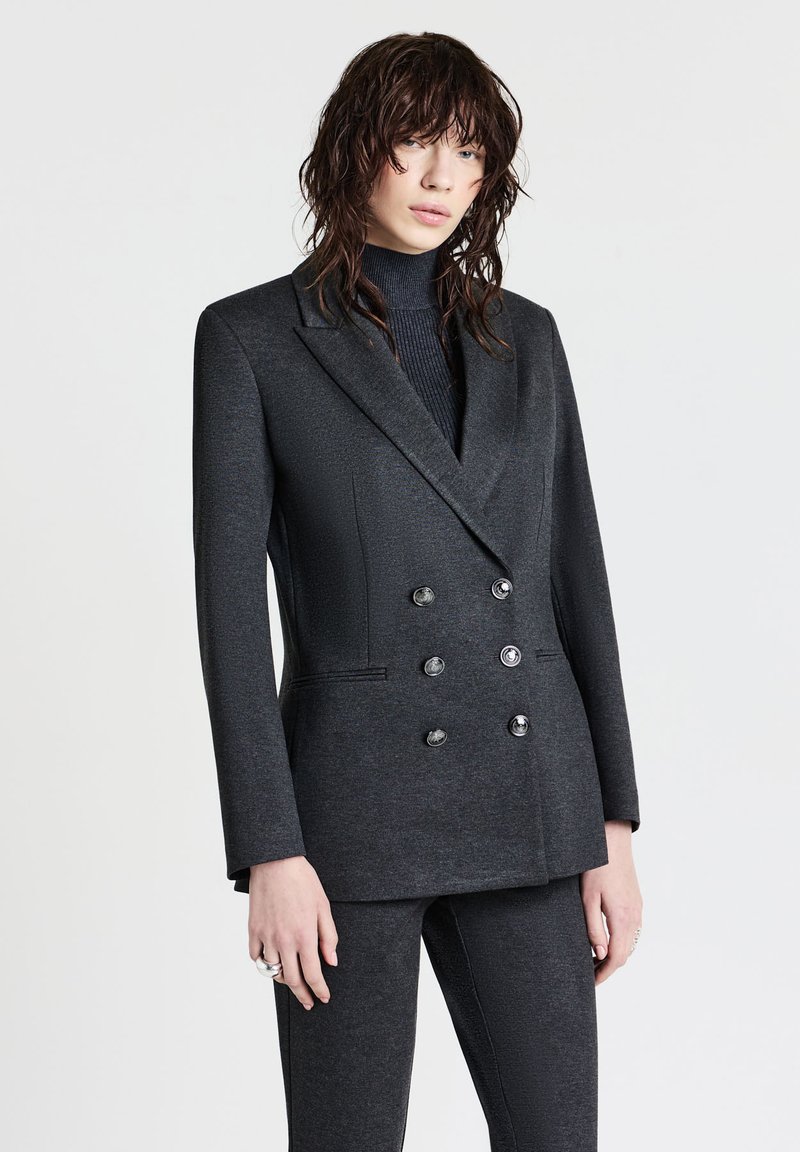 Blazer grigio scuro doppiopetto con finitura satinata, revers a lancia, sei bottoni e spalle strutturate. Abbinato a un dolcevita in maglia.