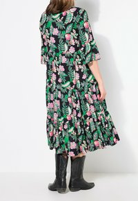 Robe noire à fleurs avec des feuilles vertes et des fleurs roses, ourlet à volants, manches courtes en forme de cloche, portée avec des bottes de cow-boy noires.