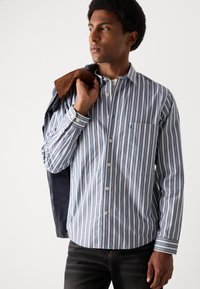 Csíkos, világoskék és fehér gombos ing sötétkék díszítéssel, hosszú ujjal és egyetlen mellzsebbel. Barna dzsekivel viselve.